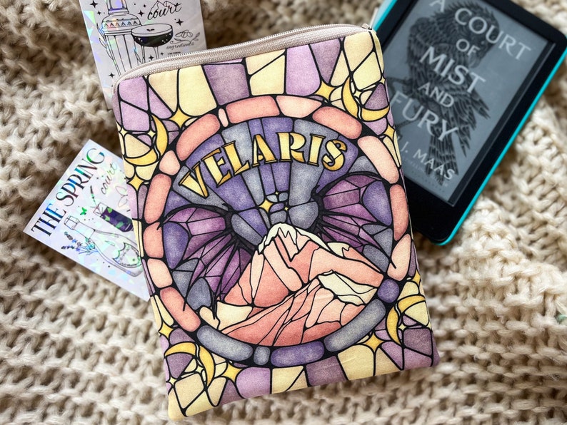 May include: A stained glass style book sleeve with a purple, pink, and yellow design. The sleeve features a mountain range and the words "VELARIS" in gold lettering.