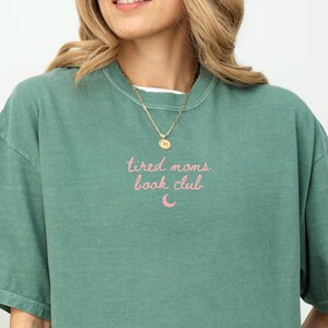 May include: A green crew neck t-shirt with the text "tired moms book club" in pink script. A gold necklace with a pendant is visible. The shirt is a solid color and has a casual style.