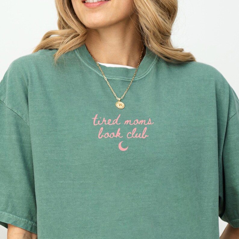 May include: A green crew neck t-shirt with the text "tired moms book club" in pink script. A gold necklace with a pendant is visible. The shirt is a solid color and has a casual style.