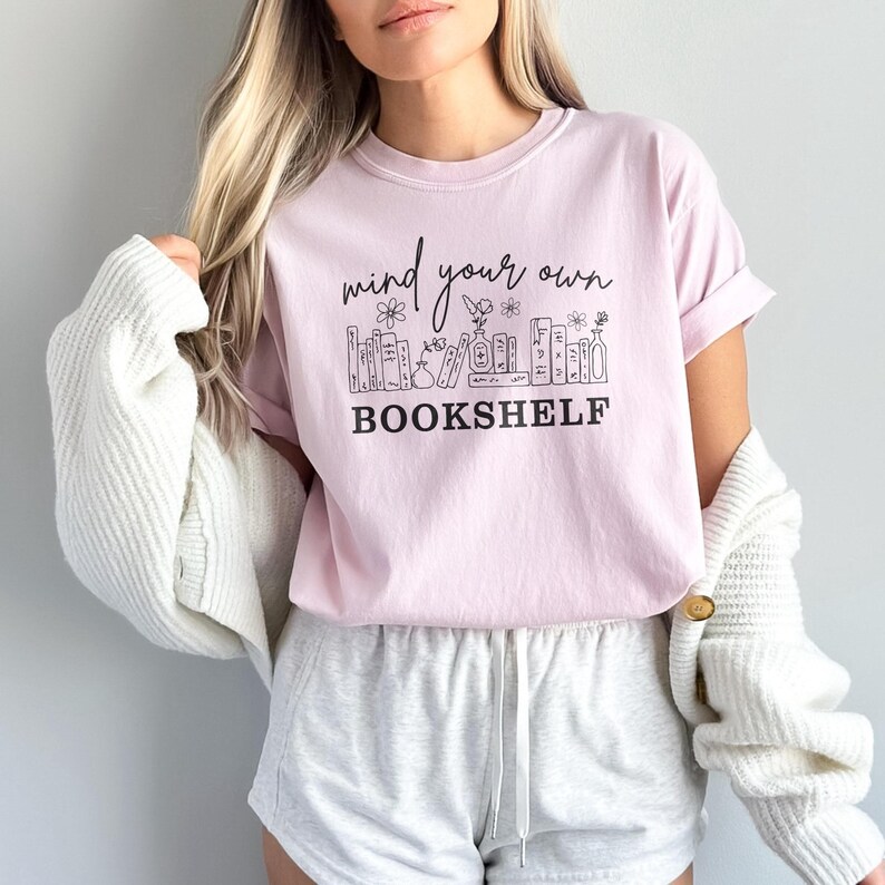 May include: A light pink t-shirt with rolled-up sleeves featuring the text "mind your own bookshelf" in black script and a bookshelf graphic. The shirt is paired with gray shorts and a white cardigan.
