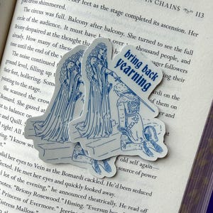 May include: Three stickers with a blue line drawing of a medieval scene. The stickers are on a book. One sticker has the text "bring back yearning". The scene depicts a kneeling figure and two figures in long gowns.