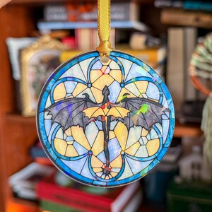 Op de afbeelding: Ronde glas-in-lood ornament met een drakenontwerp. De draak is zwart en geel, met uitgespreide vleugels. Het ornament heeft een blauw-gele rand en wordt opgehangen met een gouden lint.