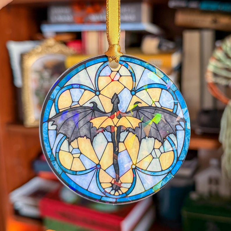 Op de afbeelding: Ronde glas-in-lood ornament met een drakenontwerp. De draak is zwart en geel, met uitgespreide vleugels. Het ornament heeft een blauw-gele rand en wordt opgehangen met een gouden lint.