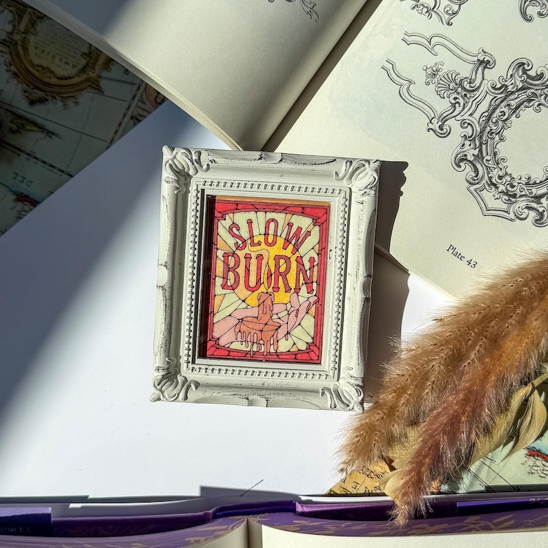 May include: A small, white framed art piece with the words "SLOW BURN" in red and yellow. The artwork depicts a candle and hands in a stained glass style. The frame has a distressed, vintage look.