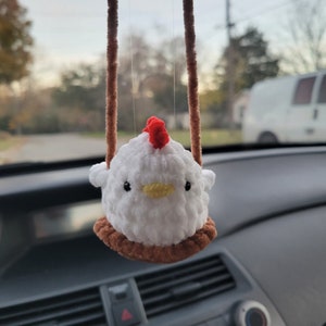 May include: A white crocheted chicken with a red comb is hanging from a brown string in a car. The chicken is on a brown swing.