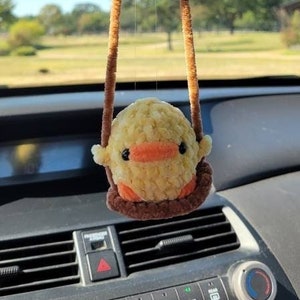 May include: A yellow and orange crocheted chick hanging from the rearview mirror of a car. The chick is sitting on a brown crocheted swing.