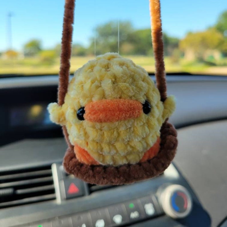 May include: A yellow crocheted duck with orange beak and black eyes hanging from a brown string. The duck is sitting on a brown crocheted swing.