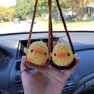 May include: Two yellow crocheted duck plushies hanging from brown strings. The ducks have orange beaks and brown eyes. The plushies are hanging from a car's rearview mirror.