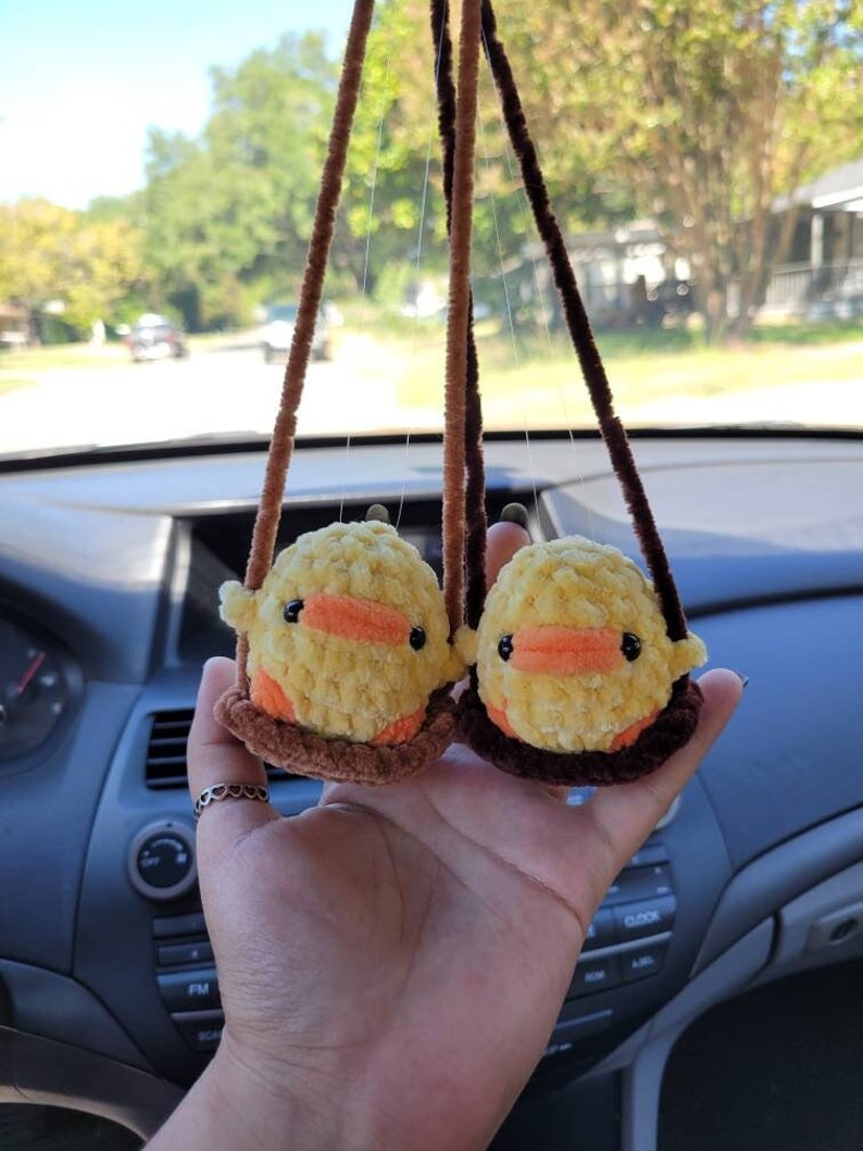 May include: Two yellow crocheted duck plushies hanging from brown strings. The ducks have orange beaks and brown eyes. The plushies are hanging from a car's rearview mirror.