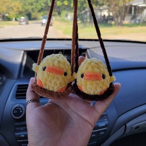 May include: Two yellow crocheted duck plushies hanging from a rearview mirror in a car. The ducks are sitting on brown crocheted swings.