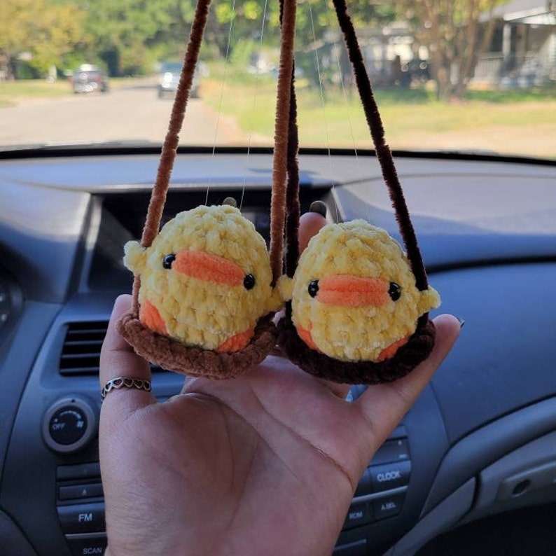 May include: Two yellow crocheted duck plushies hanging from a rearview mirror in a car. The ducks are sitting on brown crocheted swings.