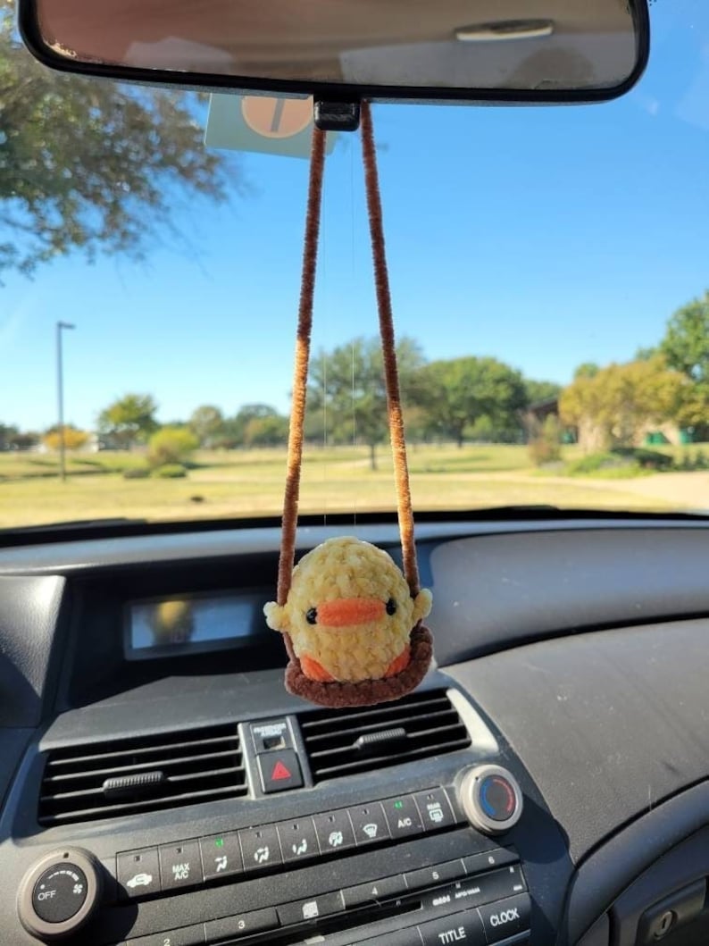 May include: A yellow and orange crocheted chick hanging from the rearview mirror of a car. The chick is sitting on a brown crocheted swing.