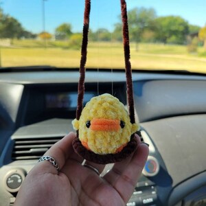 May include: A yellow and orange fuzzy chick hanging from a brown string in a car. The chick is a car charm.