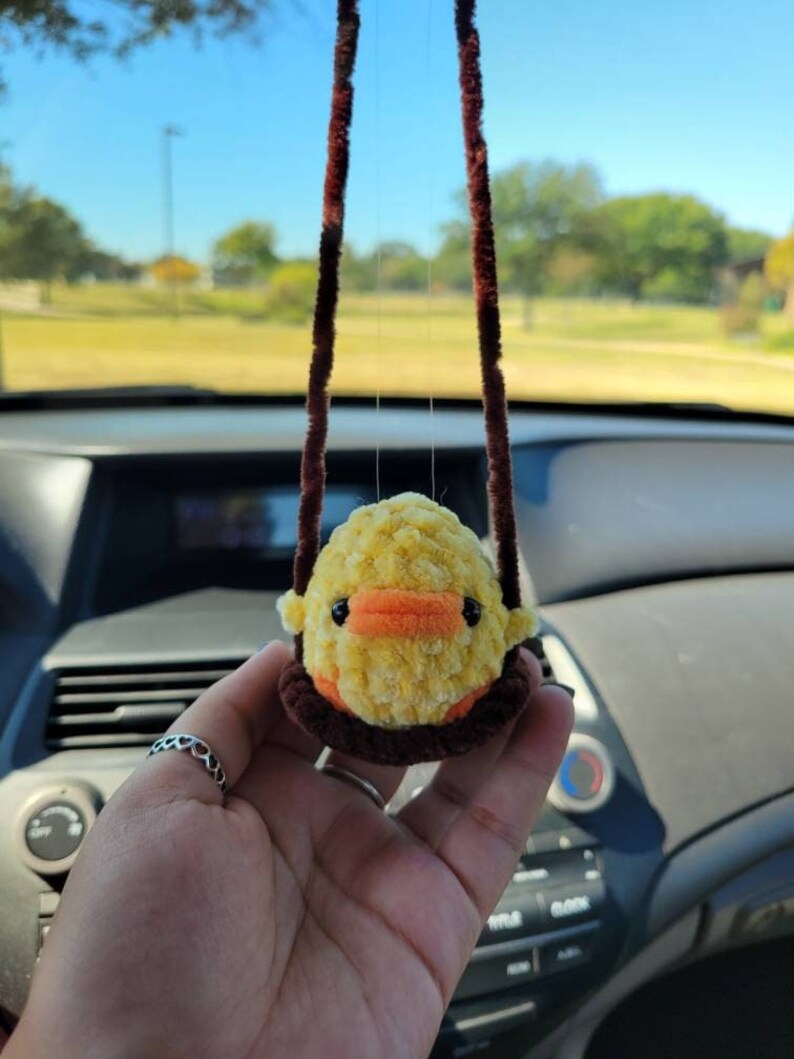 May include: A yellow and orange fuzzy chick hanging from a brown string in a car. The chick is a car charm.