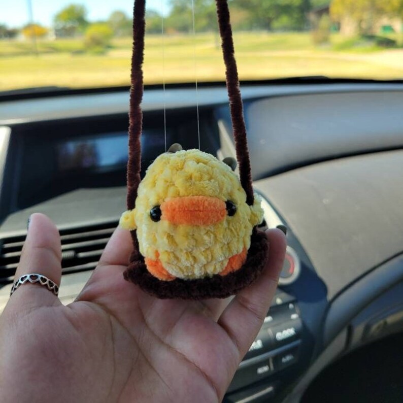 May include: A yellow crocheted chick with orange beak and brown eyes is hanging from a brown string in a car. The chick is sitting on a brown crocheted swing.