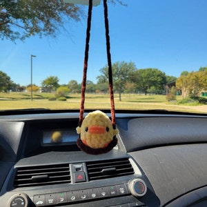 May include: A yellow duck plush toy with brown eyes and an orange beak hangs from the rearview mirror of a car. The duck is attached to a brown string.
