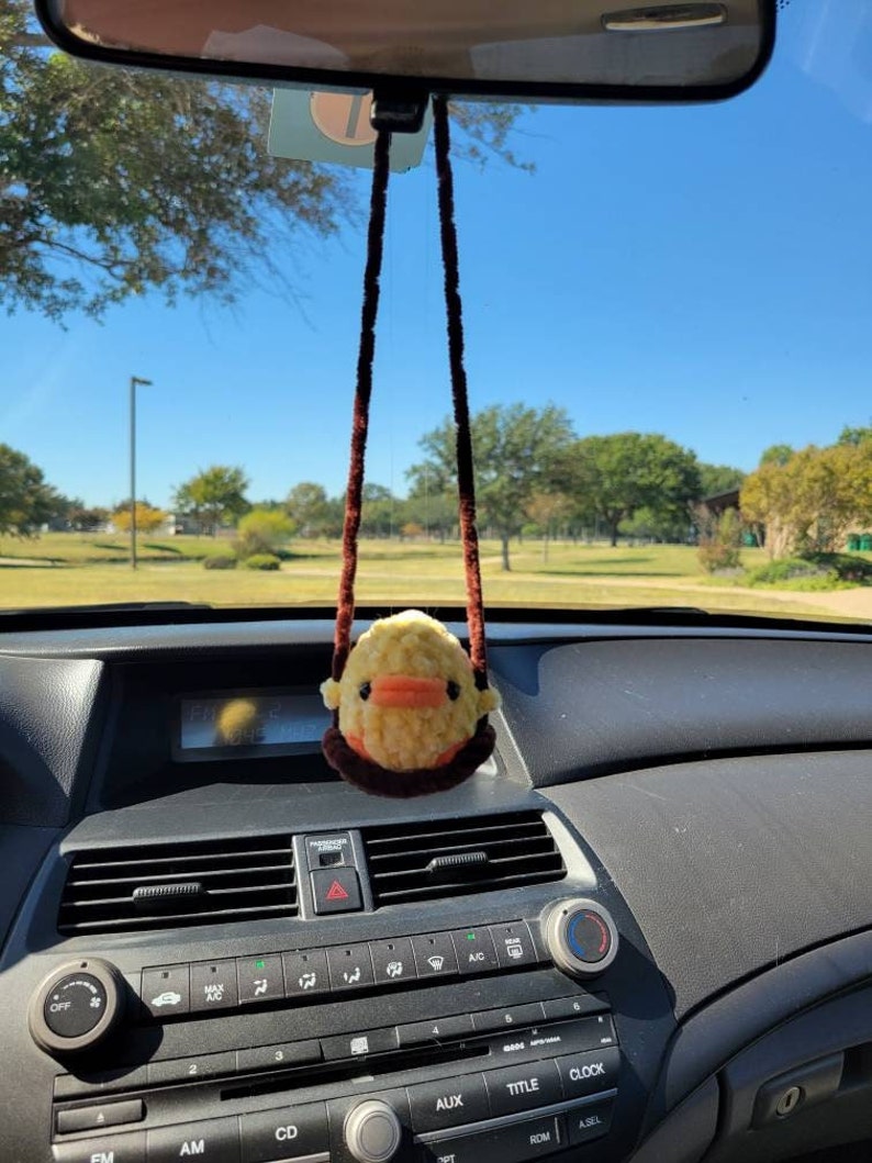 May include: A yellow duck plush toy with brown eyes and an orange beak hangs from the rearview mirror of a car. The duck is attached to a brown string.