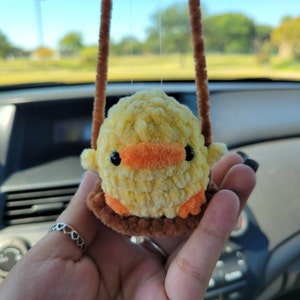May include: A yellow crocheted chick with orange beak and black eyes is hanging from a brown string. The chick is sitting on a brown crocheted swing.