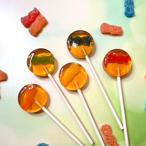 May include: Five orange lollipops with different colored gummy shapes inside. The lollipops have white sticks and are arranged on a white and green background.