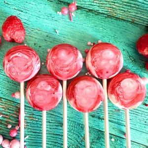 May include: Six round, red and white swirl lollipops on wooden sticks are arranged on a teal surface, alongside fresh raspberries and sprinkles. The lollipops have a marbled appearance, and the scene is brightly lit.