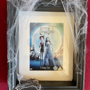 May include: A black framed picture with a white mat and a DVD cover for the movie "Corpse Bride" by Tim Burton. The cover features Johnny Depp and Helena Bonham Carter as the main characters. The frame is decorated with white spiderwebs and a black spider.