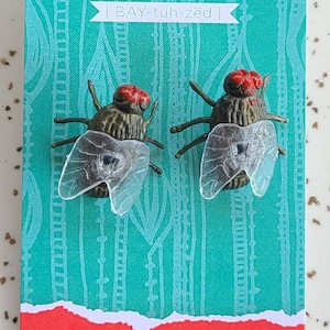 May include: A pair of fly earrings on a turquoise card with the text "Betazed Creative.com | BAY-tuh-zed | Made in Port Angeles, WA www.betazedcreative.com"