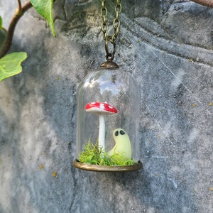 May include: A quirky pendant necklace with a small glass dome. Within the dome, a red and white mushroom and a small, glowing ghost sit on green moss. The pendant is suspended from a bronze-toned chain.