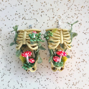 Cottagecore skeleton ribcage torso earrings with plastic greenery and five red and white hand-made polymer clay amanita muscaria mushrooms with sterling silver hooks front view