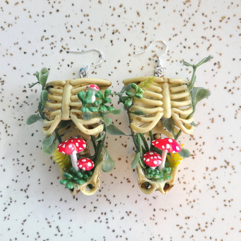 Cottagecore skeleton ribcage torso earrings with plastic greenery and five red and white hand-made polymer clay amanita muscaria mushrooms with sterling silver hooks front view