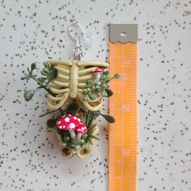 Cottagecore skeleton ribcage torso earring with plastic greenery and three red and white hand-made polymer clay amanita muscaria mushrooms with sterling silver hooks shown next to measuring tape approximately 3 inches, 7.5 cm drop length