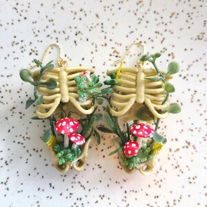Cottagecore skeleton ribcage torso earrings with plastic greenery and five red and white polymer clay amanita muscaria mushrooms with gold hooks front view
