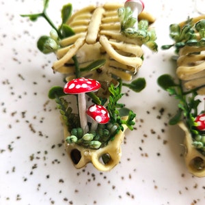 Close-up view of Cottagecore skeleton ribcage torso earrings with plastic greenery, red and white hand-made polymer clay amanita muscaria mushrooms