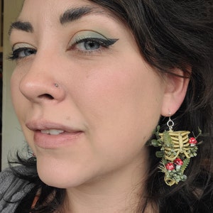 Woman wearing Cottagecore skeleton ribcage torso earrings with plastic greenery, red and white hand-made polymer clay amanita muscaria mushrooms with sterling silver hooks front view
