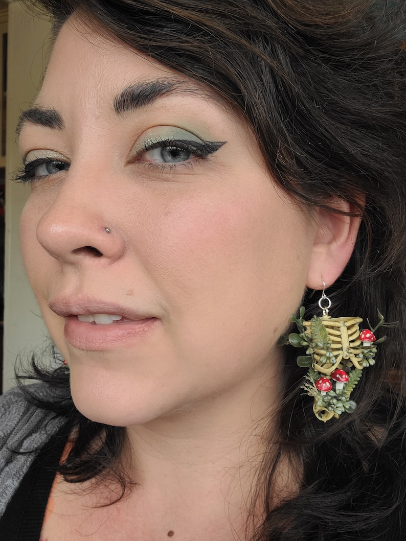 Woman wearing Cottagecore skeleton ribcage torso earrings with plastic greenery, red and white hand-made polymer clay amanita muscaria mushrooms with sterling silver hooks front view