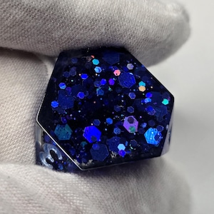 May include: A close-up of a ring with a dark blue, hexagonal face filled with iridescent glitter. The ring's band is silver-colored, and the glitter reflects various shades of blue and purple. The ring is held by a white glove.
