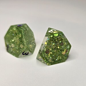 May include: Two green, multi-faceted dice with glitter and the number 3 visible. The dice are made of a clear resin and filled with various sizes of green and gold glitter. The dice are designed for tabletop games.