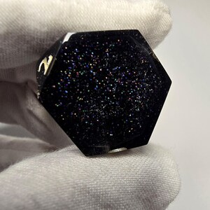 May include: A hexagonal black resin object with a flat surface embedded with fine, multi-colored iridescent glitter particles. The edges of the hexagon are smooth and dark, with a small metallic gold-colored detail visible on one side.