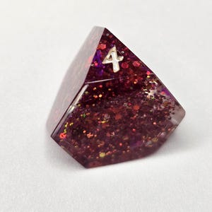 May include: A four-sided tetrahedral gaming die made of translucent dark purple resin filled with dense red, purple, and gold glitter. The top face features the number 4 engraved in a silver-colored font, clearly visible against the dark, sparkling background.