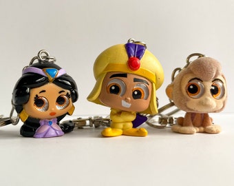 May include: Three character keychains from the movie Aladdin. The keychains feature Jasmine in a purple dress, Aladdin in yellow clothing, and Abu the monkey. Each keychain has a silver keyring attached.