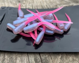 May include: A collection of soft plastic fishing lures. The lures feature a light blue body with dark speckles and vibrant pink tails. They are presented on a black surface, possibly for display or packaging.