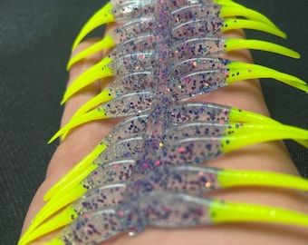 May include: A close-up shot of several clear fishing lures. Each lure has neon yellow tips and purple glitter. The lures are arranged in a row, highlighting their design and colour contrast. The image is well-lit, highlighting the details of the lures.