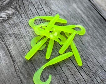 May include: A collection of neon green, soft plastic fishing lures with a curly tail design. The lures are scattered on a weathered, grey wooden surface. The lures are designed to attract fish.