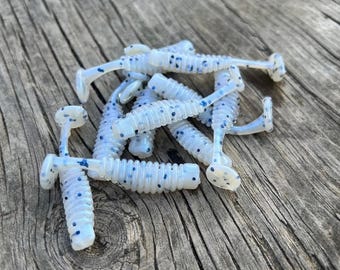 May include: A pile of soft plastic fishing lures in a pale blue hue, speckled with dark blue dots. The lures feature a ribbed body and a paddle tail. They are displayed on a weathered wooden surface.