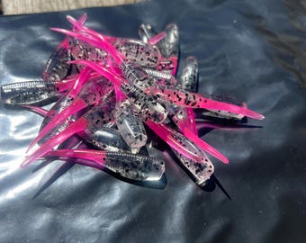 May include: A collection of soft plastic fishing lures featuring a clear, segmented body with embedded black glitter and a vibrant, solid neon pink tail. The lures are designed with a ribbed texture and a pointed, tapered end for movement.