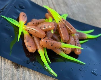 May include: A collection of small, brown fishing lures with vibrant green tails. The lures feature a speckled design and are presented on a black surface. These lures are designed to attract fish, and are likely used for angling.