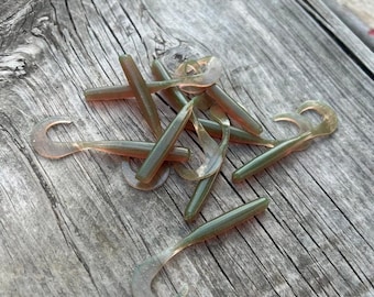 May include: A pile of soft plastic fishing lures in green and orange hues, featuring a curved tail. The lures are arranged on a weathered wooden surface, implying outdoor use and angling.