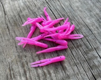 May include: A collection of vibrant pink soft plastic fishing lures with a forked tail design. The lures are arranged on a weathered, grey wooden surface. Each lure features small black speck details.