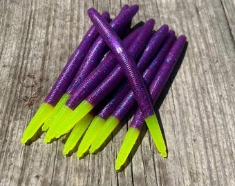 May include: A collection of purple fishing lures with a shimmering finish and vivid neon yellow tips. The lures are displayed on a weathered wooden surface, highlighting their bright colours and textured design.