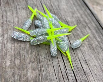 May include: A collection of silver glitter fishing lures with vibrant neon green tails. The lures are displayed on a weathered wooden surface, contrasting the shiny lures with the rustic background.