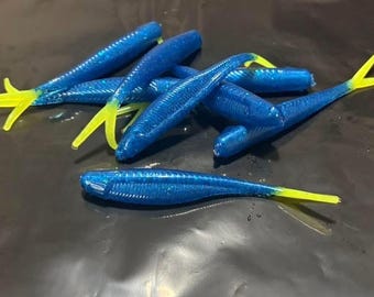 May include: A collection of soft plastic fishing lures featuring a blue glitter body with a segmented texture and a bright yellow split tail. These lures are designed with a streamlined, fish-like shape and are presented in a group.